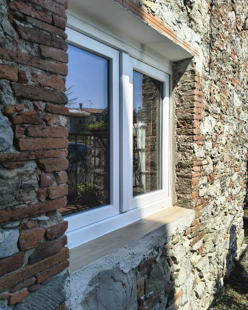 Finestra bianca a due ante in PVC montata in un muro di pietra e mattoni, vista dall’esterno, con davanzale in legno